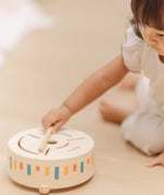 Close up of a child hand placing the wooden drumstick on top of the PlanToys wooden sensory drum musical toy.