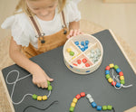 Overhead view of child playing with the PlanToys threading beads wooden lacing toy. Rainbow coloured wooden beads with black and white laces in a handy wooden storage box with compartments placed on a Plan Toys child's desk.