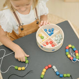 Overhead view of child playing with the PlanToys threading beads wooden lacing toy. Rainbow coloured wooden beads with black and white laces in a handy wooden storage box with compartments placed on a Plan Toys child's desk.