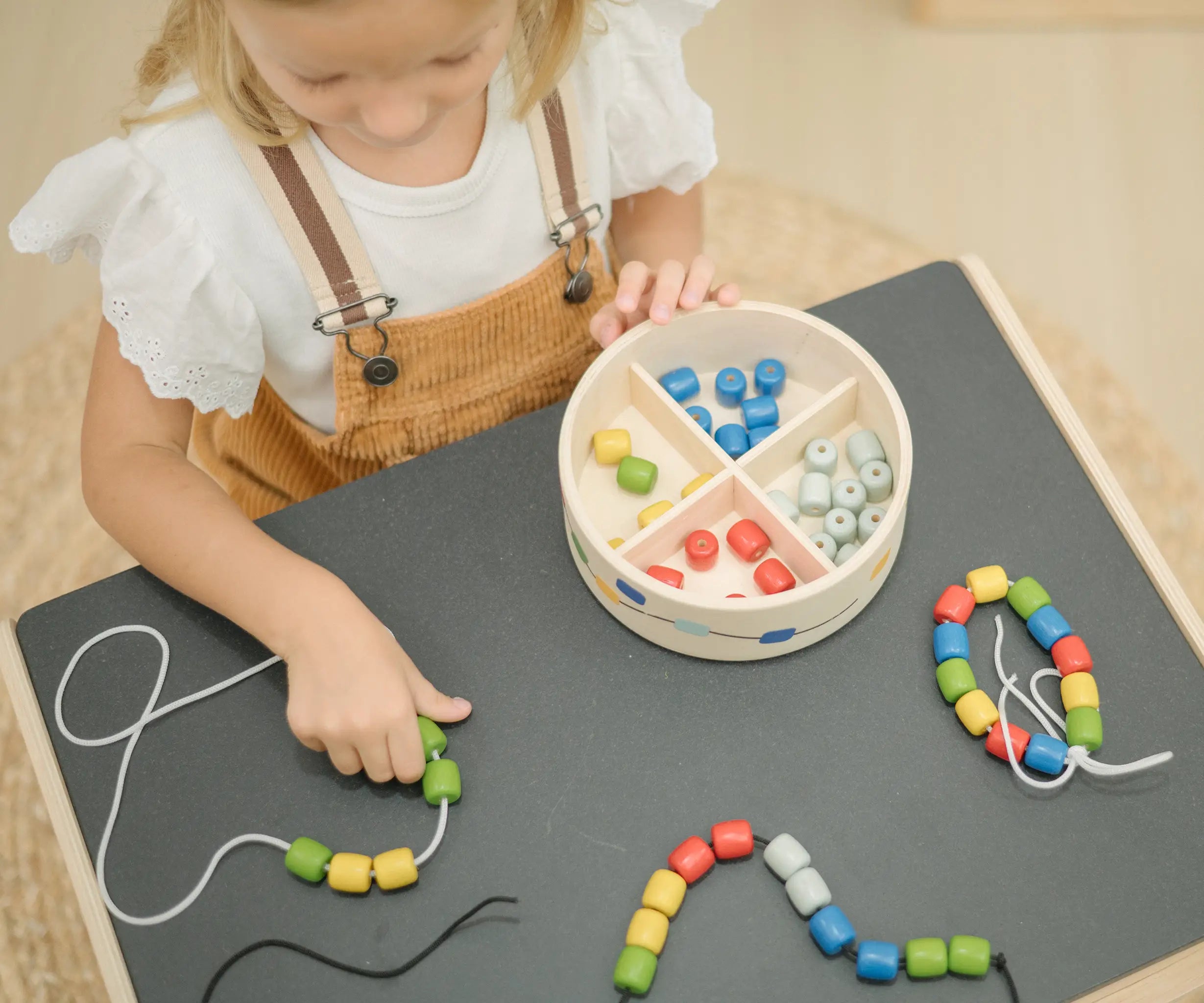 Overhead view of child playing with the PlanToys threading beads wooden lacing toy. Rainbow coloured wooden beads with black and white laces in a handy wooden storage box with compartments placed on a Plan Toys child's desk.