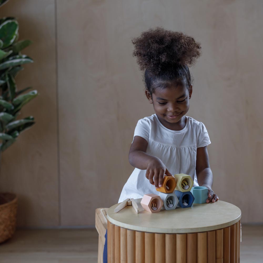 The PlanToys Bee Hives in the Orchard colour way placed side table. A child can be seen standing behind the table holding on to the orange coloured bee hive with a bee inside. 