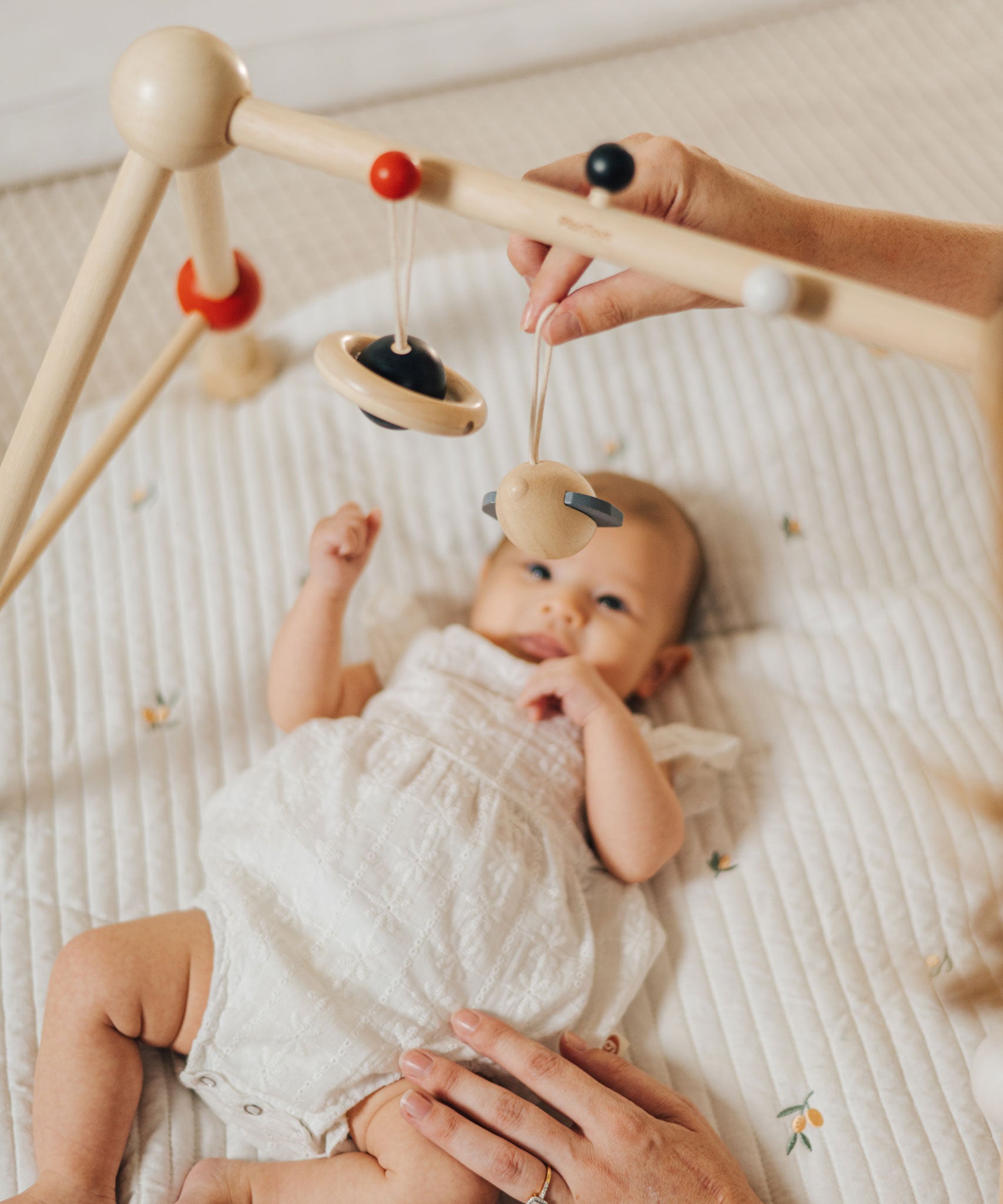 A baby laying underneath the PlanToys High Contrast Wooden Baby Play Gym looking up at the high contrast hanging toys as a. This wooden baby gym has high contrast coloured features and detailing. The gym comes with two wooden hanging toys, a planet and a rocket. PlanToys range of toys are made from sustainably sourced rubberwood, we're proud stockists of PlanToys here at Babipur.  adult placed one of the toys on the gym