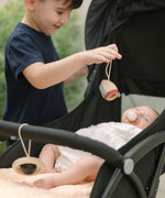A young child standing near a pushchair holding up the rocket shaped wooden toy from the PlanToys High Contrast Wooden Baby Play Gym with a baby laying underneath . The wooden baby gym has high contrast coloured features and detailing. The gym comes with two wooden hanging toys, a planet and a rocket. PlanToys range of toys are made from sustainably sourced rubberwood, we're proud stockists of PlanToys here at Babipur. 