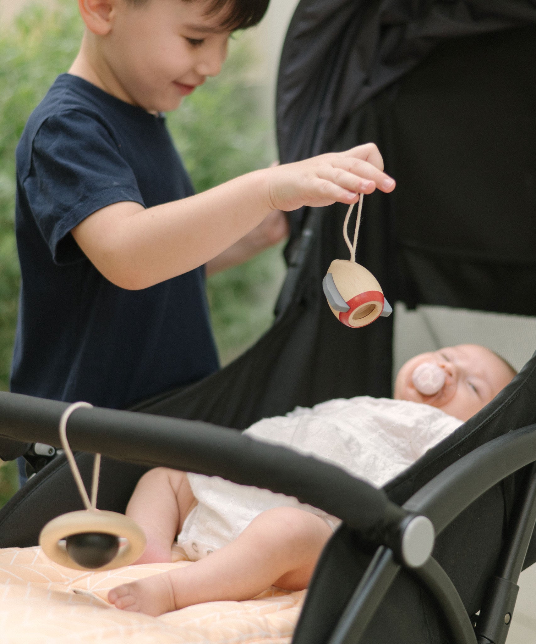 A young child standing near a pushchair holding up the rocket shaped wooden toy from the PlanToys High Contrast Wooden Baby Play Gym with a baby laying underneath . The wooden baby gym has high contrast coloured features and detailing. The gym comes with two wooden hanging toys, a planet and a rocket. PlanToys range of toys are made from sustainably sourced rubberwood, we're proud stockists of PlanToys here at Babipur. 