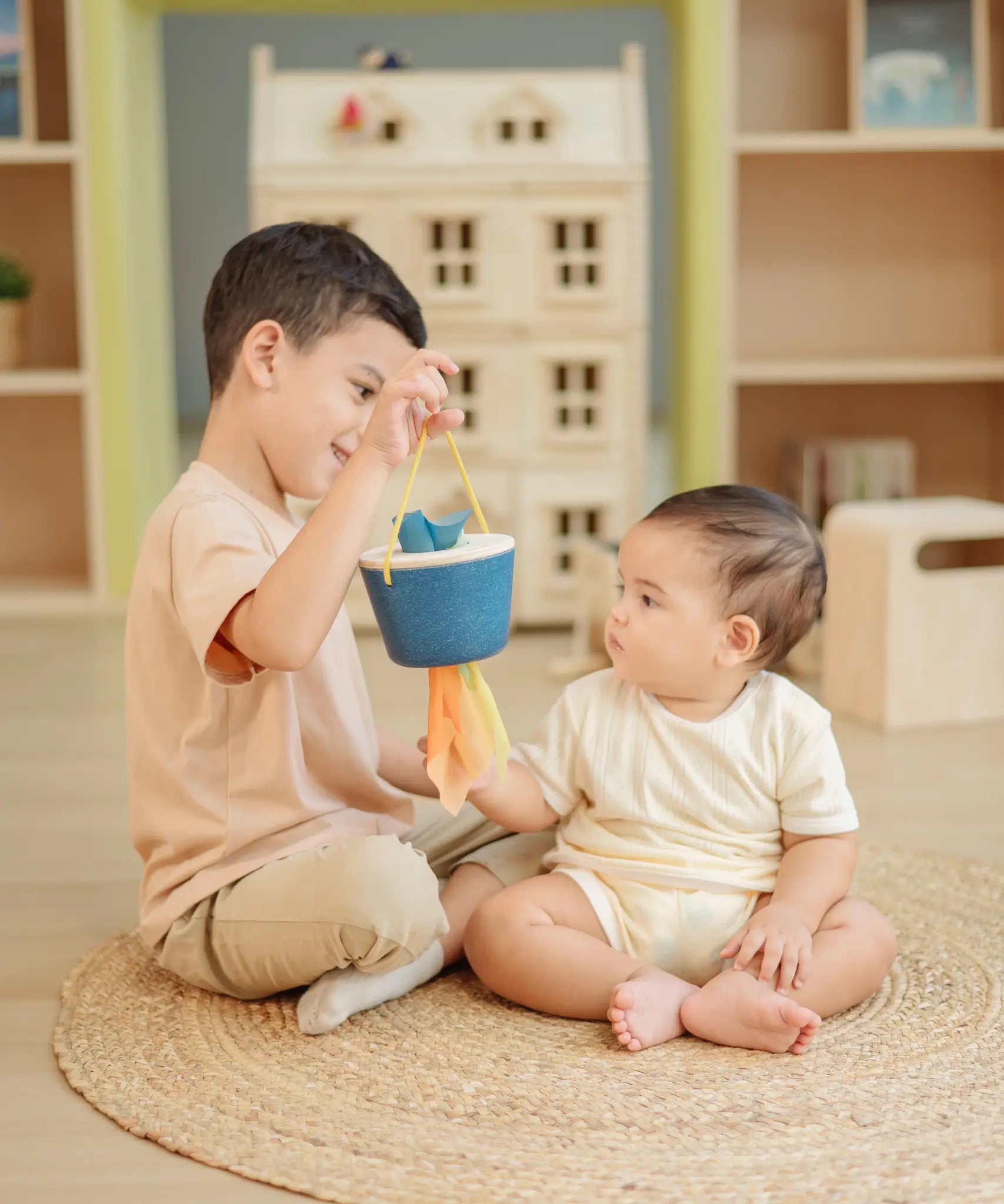 Child holding the PlanToys baby tissue box wooden sensory toy in front of a baby whilst the baby reaches for the material sticking out.