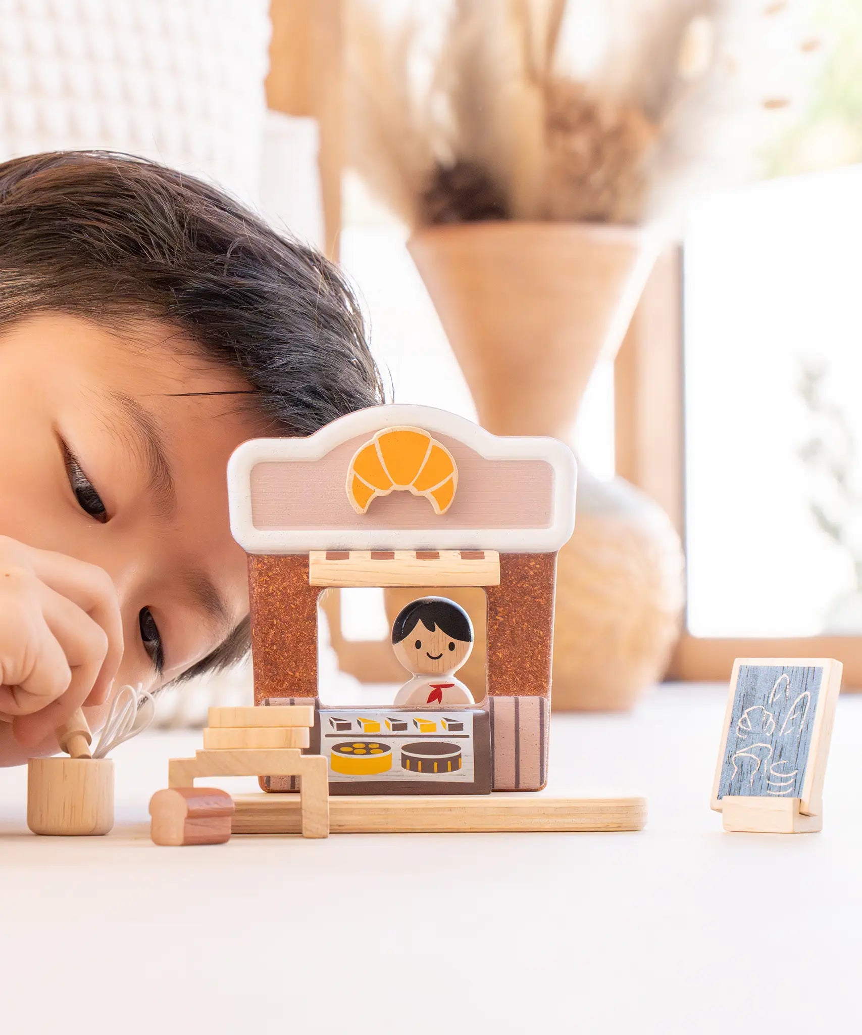 Child playing with the PlanToys bakery shop wooden toy playset, placing baking tools into a pot.