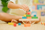 Child placing blocks on the PlanToys balancing cargo wooden balance game. Stack of wooden blocks balanced on a base piece placed on an upturned arch piece.