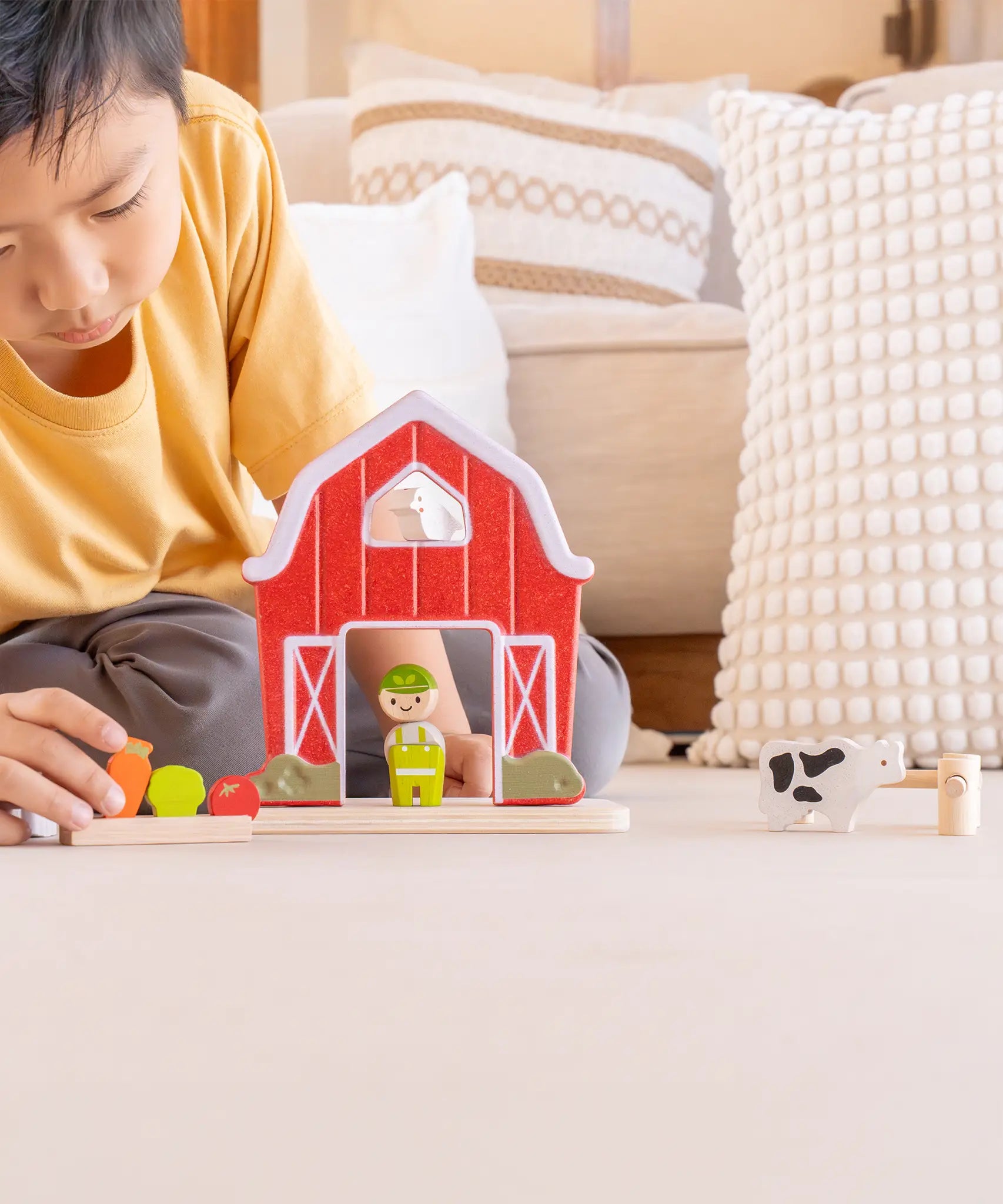 Child playing with the PlanToys barn house small world play set. Red wooden barn, mini veg patch, fences, cow, sheep, hen and farmer figure. 