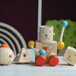 The PlanToys Build a Robot placed on a wooden surface n front of other wooden toys. The robot has it's cylinder shaped head on with a sad expression. 