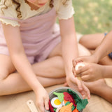 A PlanToys chef salad bowl filled with play food on a hessian blanket with children sitting 