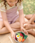 A PlanToys chef salad bowl filled with play food on a hessian blanket with children sitting 