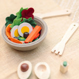 A PlanToys wooden salad bowl filled with play food next to wooden cutlery and a loose avocado play food piece