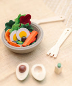 A PlanToys wooden salad bowl filled with play food next to wooden cutlery and a loose avocado play food piece