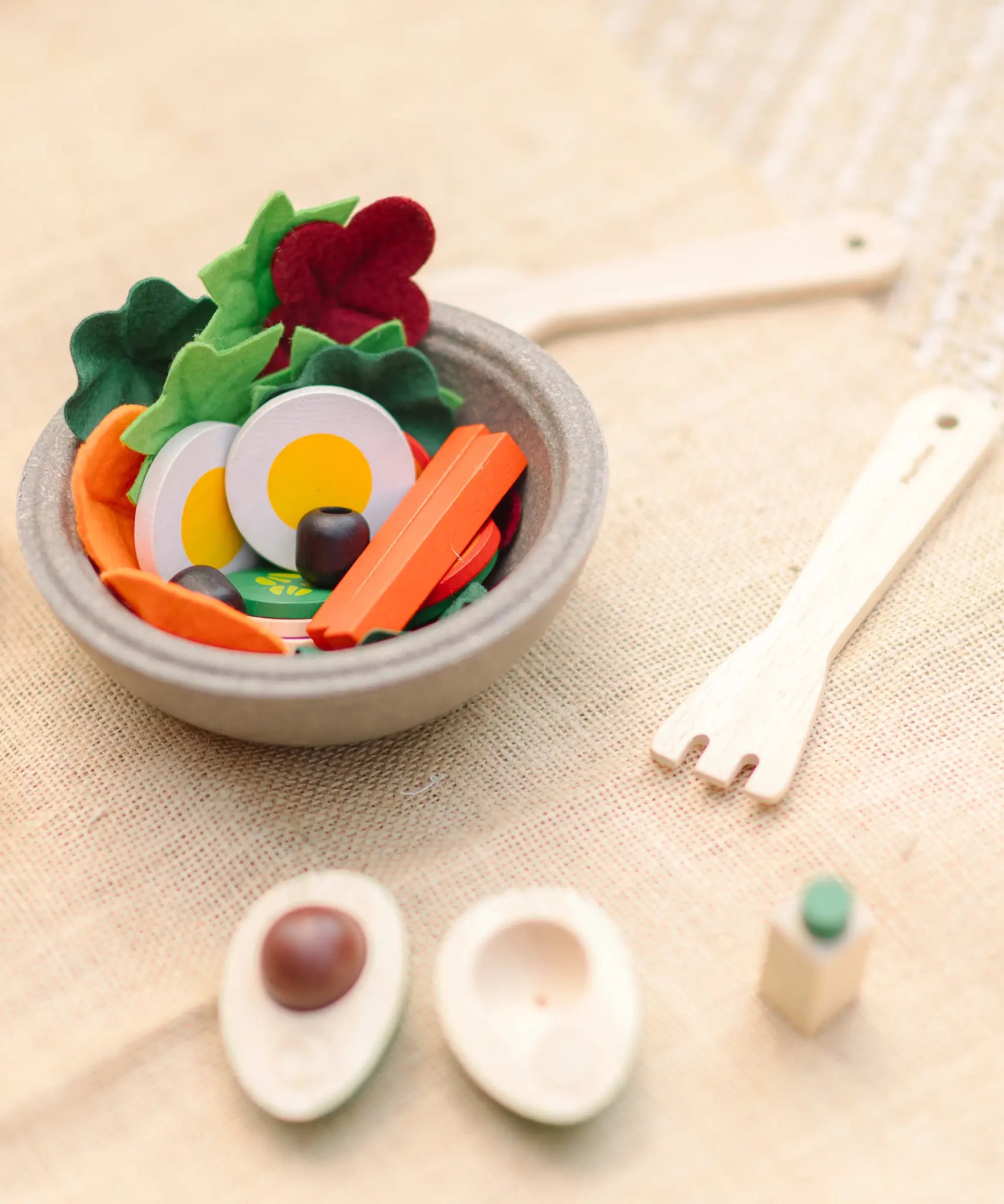 A PlanToys wooden salad bowl filled with play food next to wooden cutlery and a loose avocado play food piece