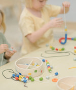 Children threading beads onto laces from the PlanToys threading beads wooden lacing toy. The wooden storage box is in front of them.
