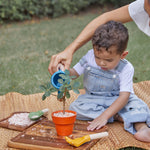 A child playing with the PlanToys Gardening Set, an adult is sitting behind them helping them use the watering can. 