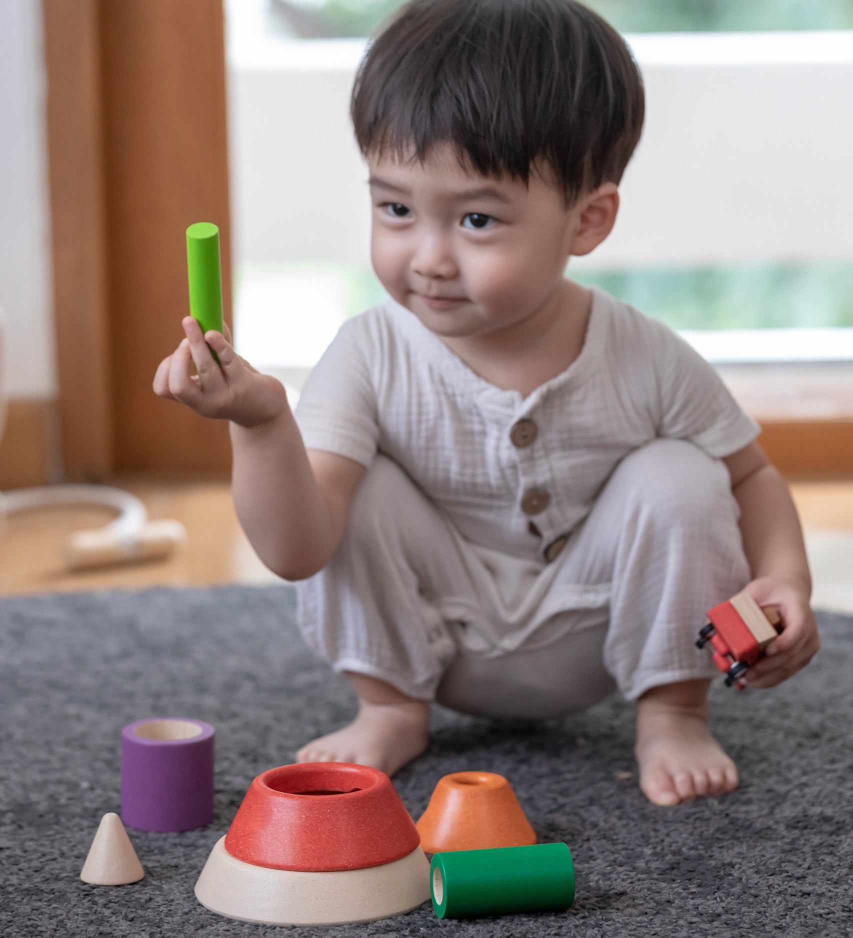 A child sitting on a grey coloured rug playing with the PlanToys Coloured Sorting Cones. 
