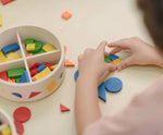 Close up of child's hand placing pieces from the PlanToys mosaic set into a circle shape. Brightly coloured wooden pieces in a storage box.