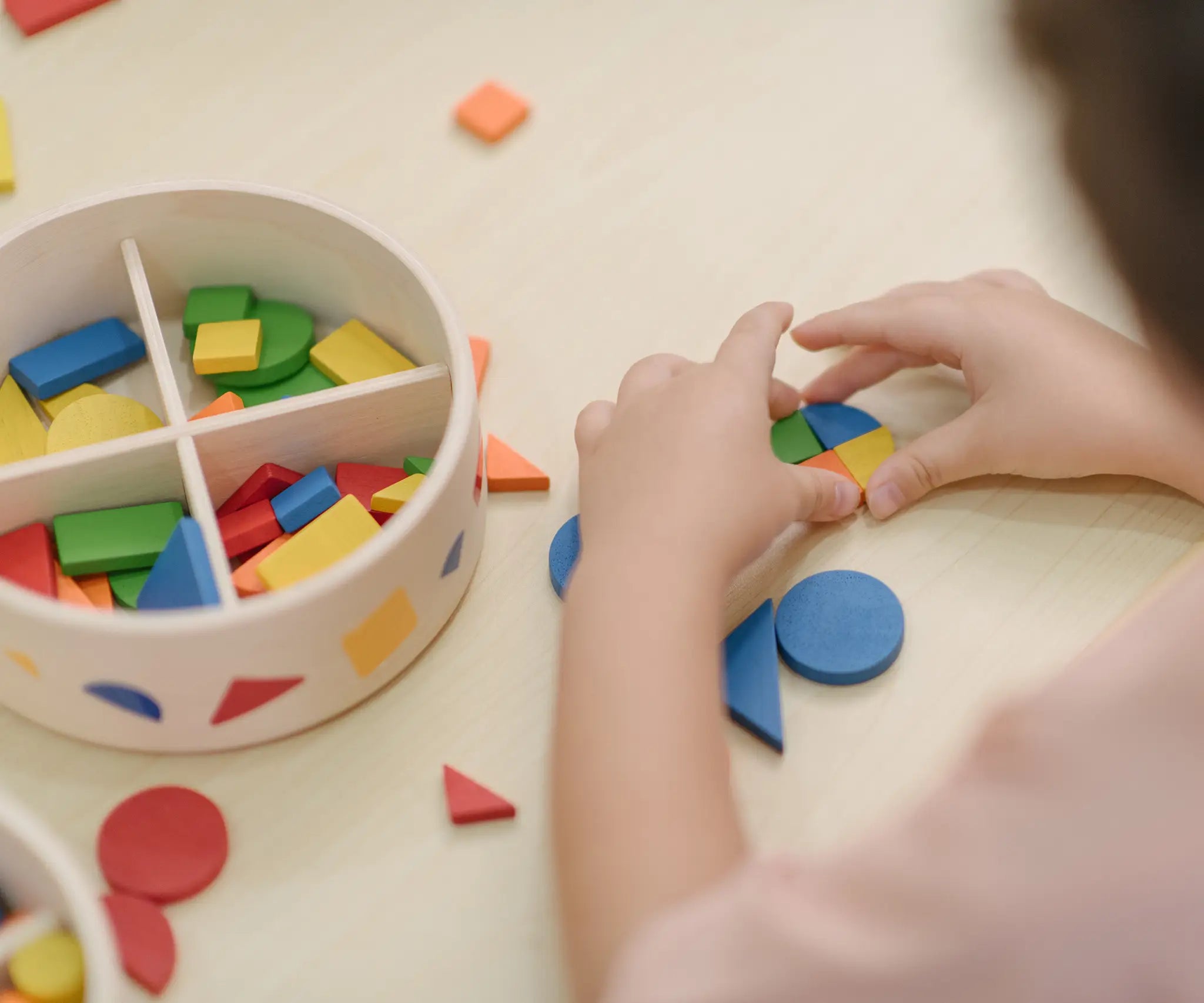 Close up of child's hand placing pieces from the PlanToys mosaic set into a circle shape. Brightly coloured wooden pieces in a storage box.
