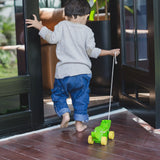 Toddler pulling the Plan Toys Dancing Alligator wooden pull along toy through a doorway onto a wooden deck