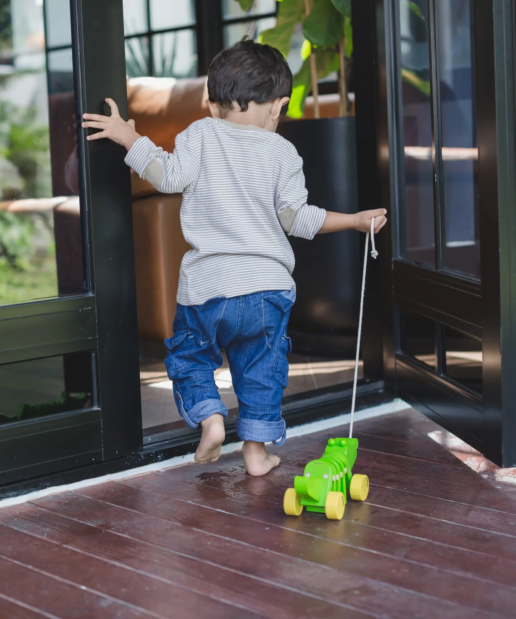 Toddler pulling the Plan Toys Dancing Alligator wooden pull along toy through a doorway onto a wooden deck