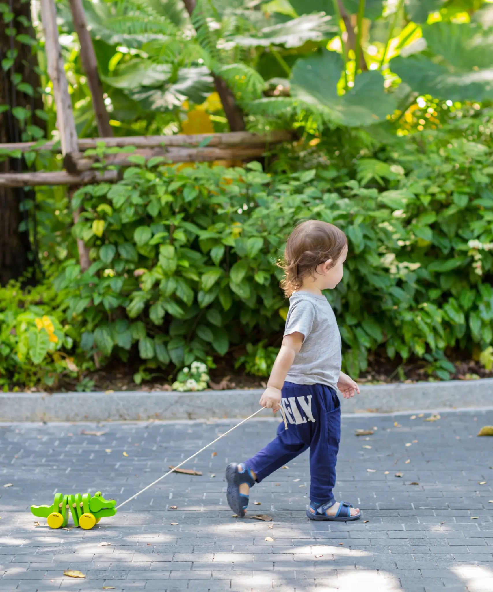 Toddler pulling the Plan Toys Dancing Alligator wooden pull along toy along an outdoor garden path