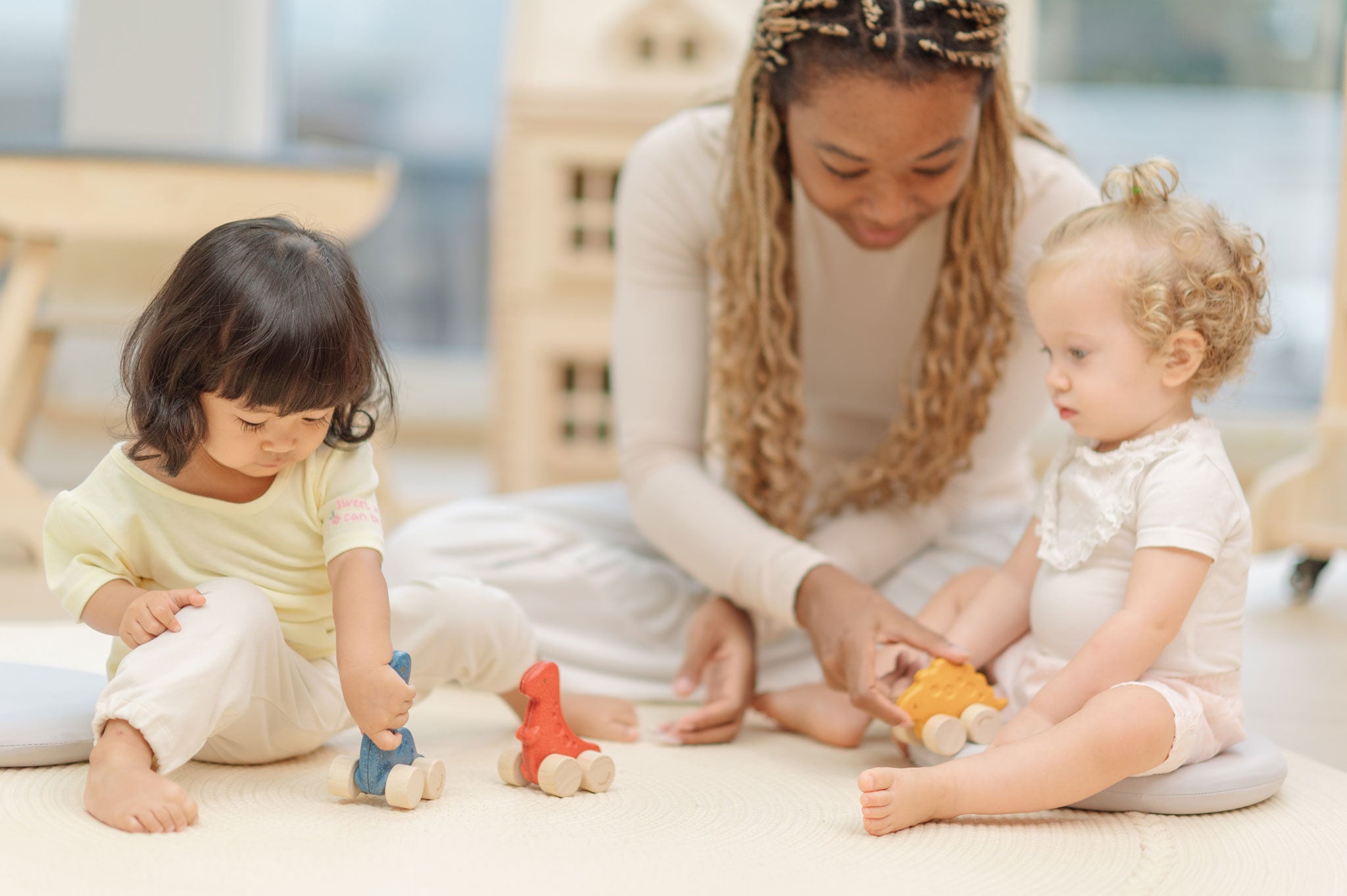 Two children and an adult playing with the PlanToys Dino wooden Car toys. A blue coloured diplodocus, a yellow stegosaurus and red coloured t-rex dinosaur figures on wheels. PlanToys range of toys are made from sustainably sourced rubberwood, we're proud stockists of PlanToys here at Babipur. 