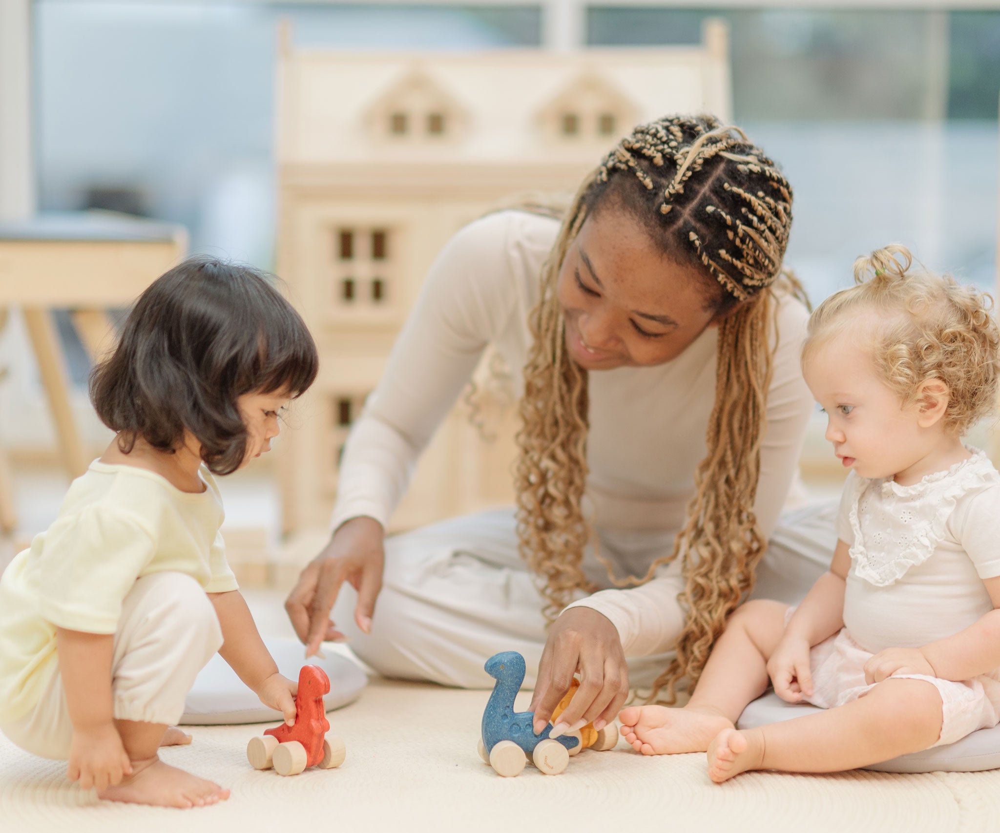 Two children and an adult playing with the PlanToys Dino wooden Car toys. A blue coloured diplodocus, a yellow stegosaurus and red coloured t-rex dinosaur figures on wheels. PlanToys range of toys are made from sustainably sourced rubberwood, we're proud stockists of PlanToys here at Babipur. 