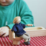 A dad doll from the PlanToys Dolls House Family with White Skin, Blonde Hair placed in a seated position on a little PlanToys wooden toilet. 
