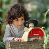 Achild standing behind the PlanToys Driving Console wooden toy placed on a wooden surface outdoors.