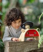 Achild standing behind the PlanToys Driving Console wooden toy placed on a wooden surface outdoors.