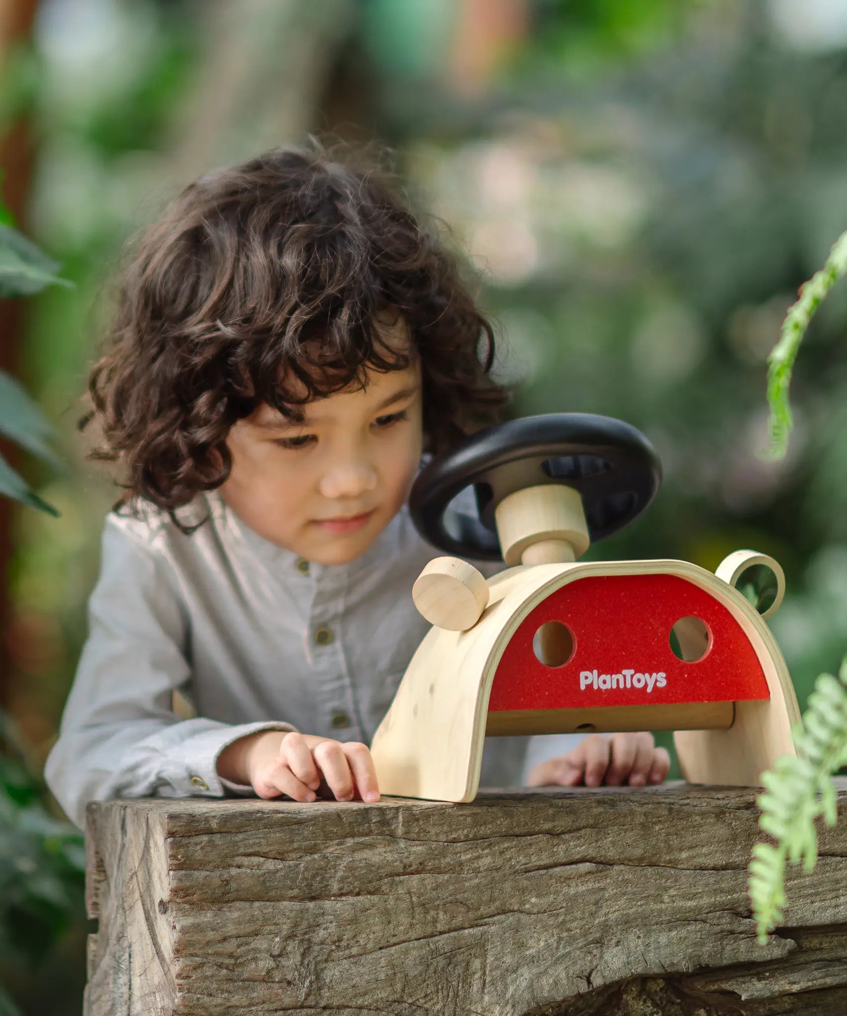Achild standing behind the PlanToys Driving Console wooden toy placed on a wooden surface outdoors.
