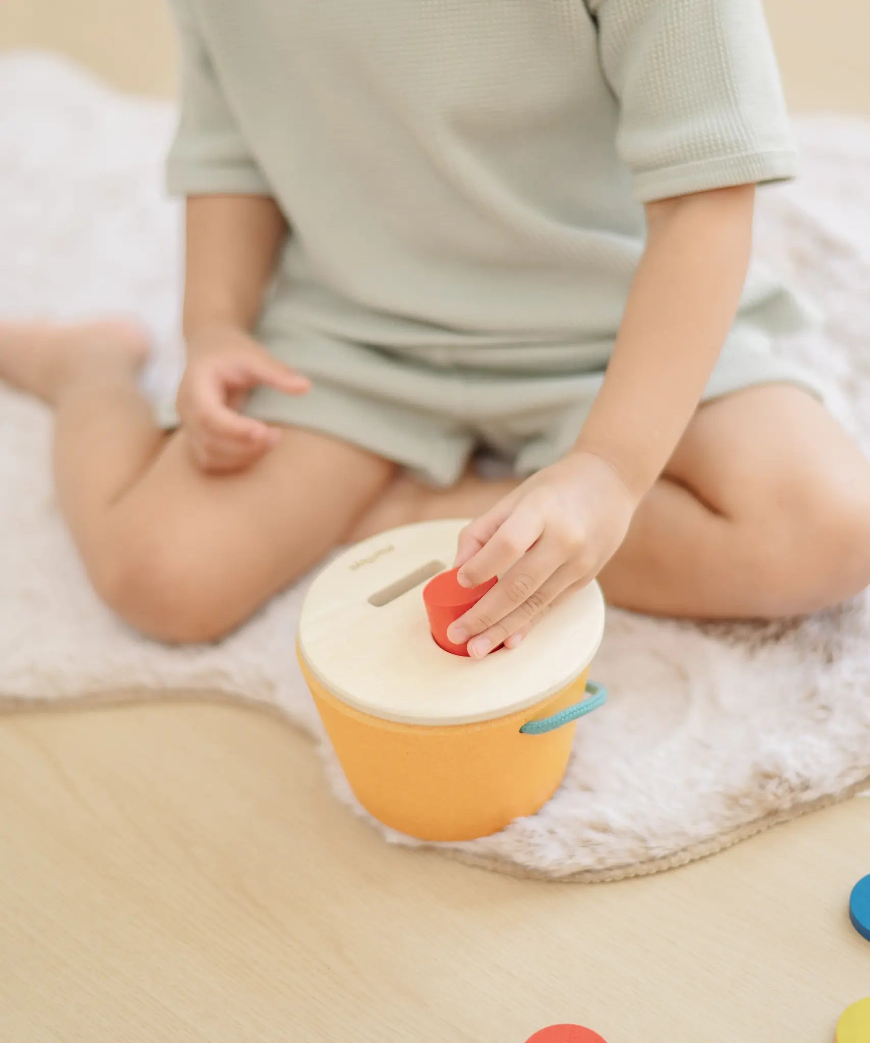 Child playing with the PlanToys drop and fit bucket wooden object permanence toy placing a red cylinder into the slot on the top of the bucket .