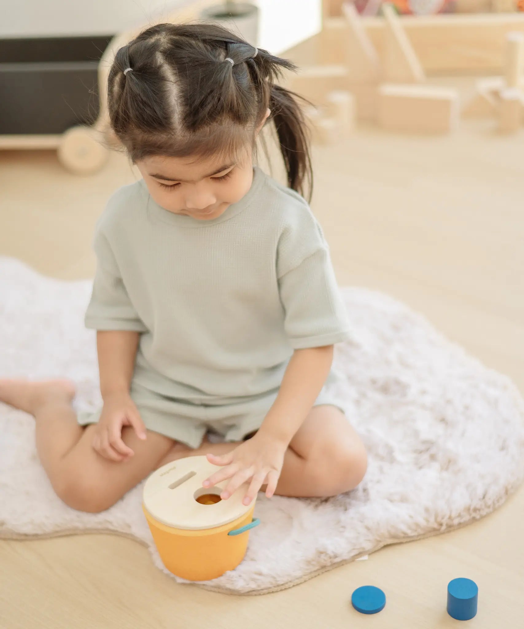 Child playing with the PlanToys drop and fit bucket wooden object permanence toy placing a red cylinder into the slot on the top of the bucket .