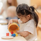 Child playing with the PlanToys drop and fit bucket wooden object permanence toy pouring the cylinder blocks and coloured coins out of the bucket .