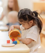 Child playing with the PlanToys drop and fit bucket wooden object permanence toy pouring the cylinder blocks and coloured coins out of the bucket .