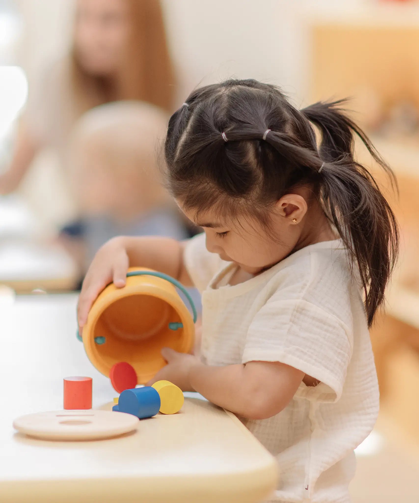 Child playing with the PlanToys drop and fit bucket wooden object permanence toy pouring the cylinder blocks and coloured coins out of the bucket .