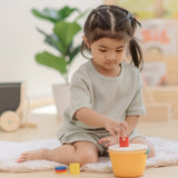 Child playing with the PlanToys drop and fit bucket wooden object permanence toy placing a red cylinder into the slot on the top of the bucket .