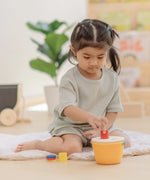 Child playing with the PlanToys drop and fit bucket wooden object permanence toy placing a red cylinder into the slot on the top of the bucket .