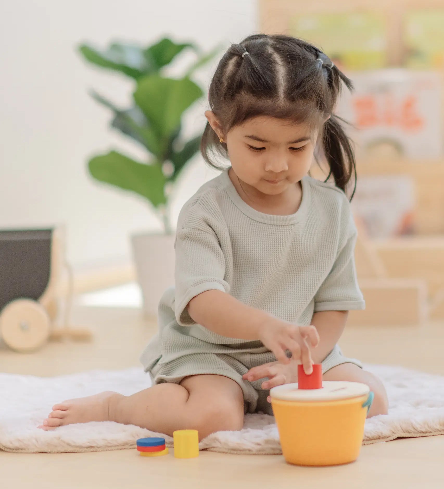 Child playing with the PlanToys drop and fit bucket wooden object permanence toy placing a red cylinder into the slot on the top of the bucket .