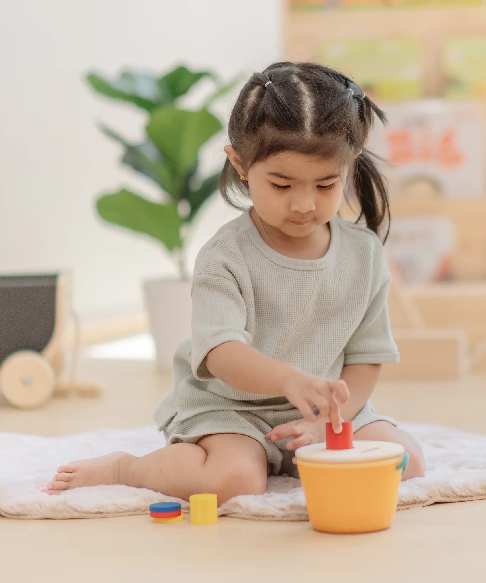 Child playing with the PlanToys drop and fit bucket wooden object permanence toy placing a red cylinder into the slot on the top of the bucket .
