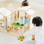 Two children playing with the PlanToys Green Dolls' House and Furniture Set. The house is placed on a white coloured rug and the children are sitting on opposite sides of the house. 