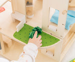 A close up of the grass effect painted areas on the side of the PlanToys Green Dolls' House and Furniture Set. A child's hand can be seen reaching for a green grass like pom pom. 