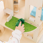 A close up of the grass effect painted areas on the side of the PlanToys Green Dolls' House and Furniture Set. A child's hand can be seen reaching for a green grass like pom pom. 