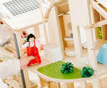 A close up of the grass effect painted areas on the side of the PlanToys Green Dolls' House and Furniture Set. A child's hand can be seen besides the grassy area holding a doll wearing a red outfit. 