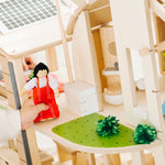 A close up of the grass effect painted areas on the side of the PlanToys Green Dolls' House and Furniture Set. A child's hand can be seen besides the grassy area holding a doll wearing a red outfit. 