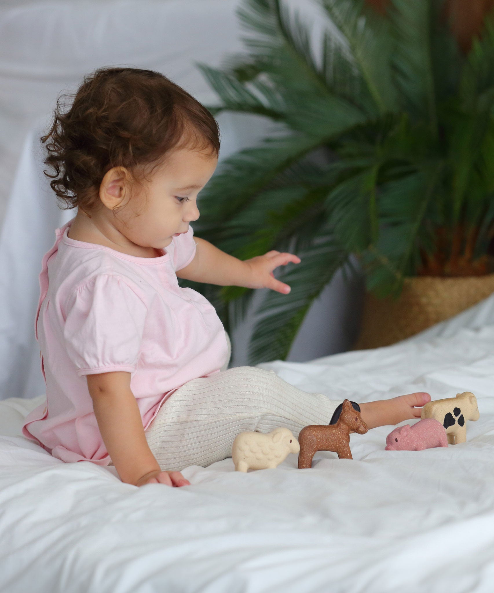 A child sitting on a white duvet playing with the figures from the  PlanToys Farm Animals Set. 
