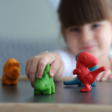 The PlanToys Dino Set on a PlanToys table, a child's hand is on the green dinosaur and the child can be seen in the background.