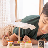 Child playing with the PlanToys Farmer's Market wooden toy playset. The child is placing produce on the wooden toy scales with the worker figure.