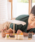 Child playing with the PlanToys Farmer's Market wooden toy playset. The child is placing produce on the wooden toy scales with the worker figure.